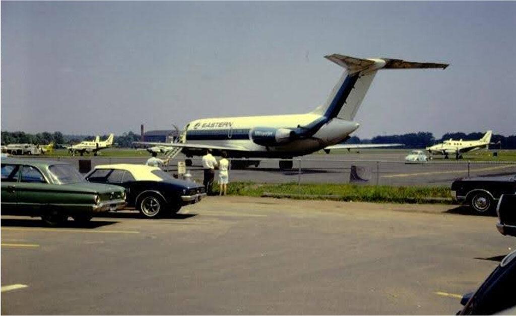 1970s - Photograph of a parking lot in the foreground and a Eastern Air Lines airplane on a runway in the background